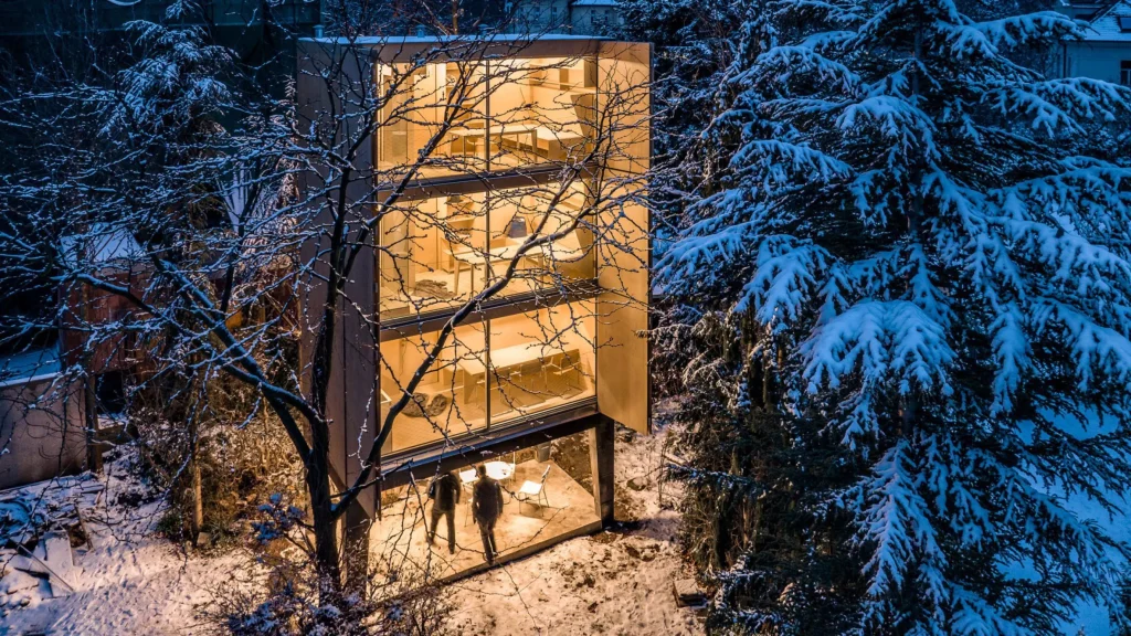 Night view of the glowing three-storey Wardrobe House with open shutters, people under the raised base, and snow-covered trees