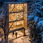Night view of the glowing three-storey Wardrobe House with open shutters, people under the raised base, and snow-covered trees