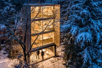 Night view of the glowing three-storey Wardrobe House with open shutters, people under the raised base, and snow-covered trees