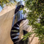 Black steel spiral staircase rising against the plywood facade amid green leaves