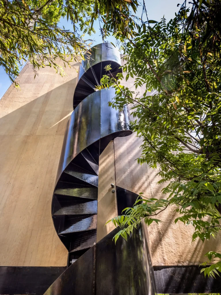 Black steel spiral staircase rising against the plywood facade amid green leaves