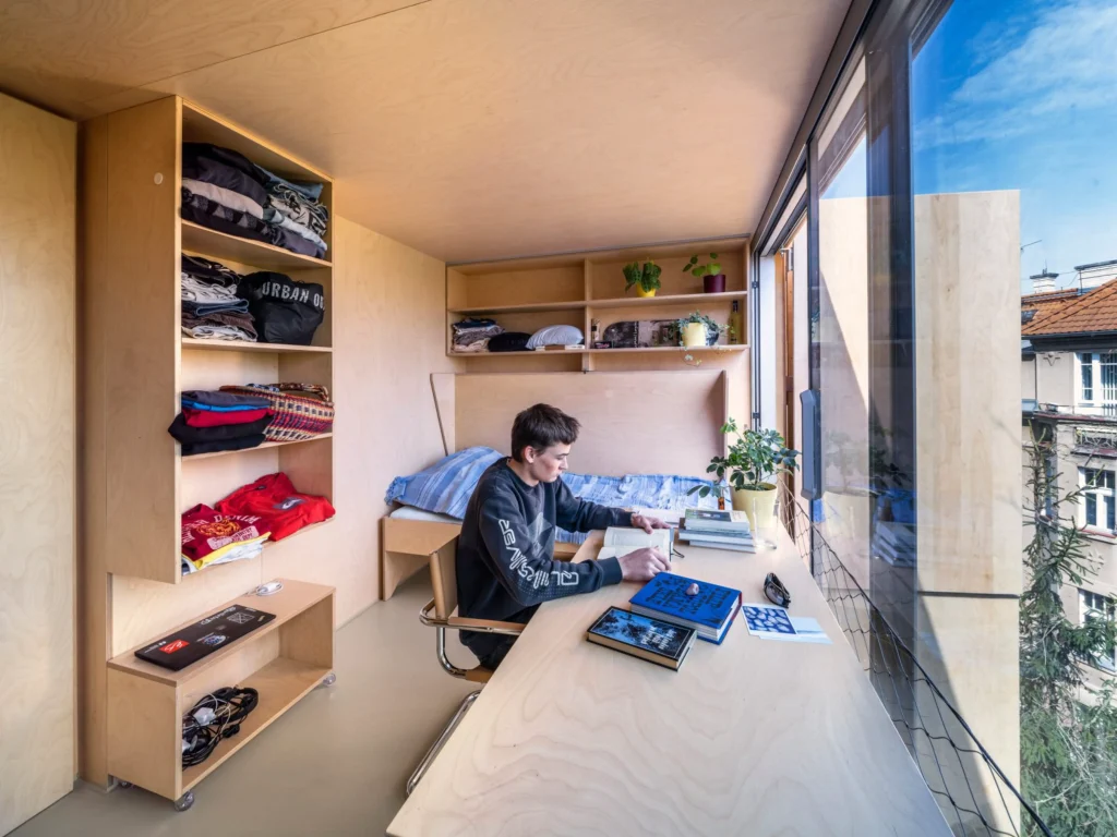 Bedroom interior with open plywood shelving, desk, fold-down bed and a young person studying by the window