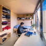Bedroom interior with open plywood shelving, desk, fold-down bed and a young person studying by the window