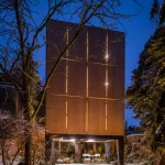 Night exterior of the closed plywood facade with vertical light lines and illuminated patio below