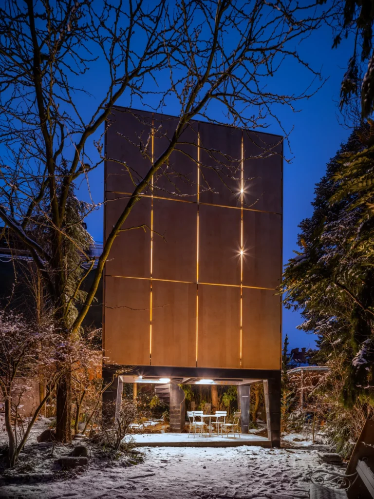 Night exterior of the closed plywood facade with vertical light lines and illuminated patio below