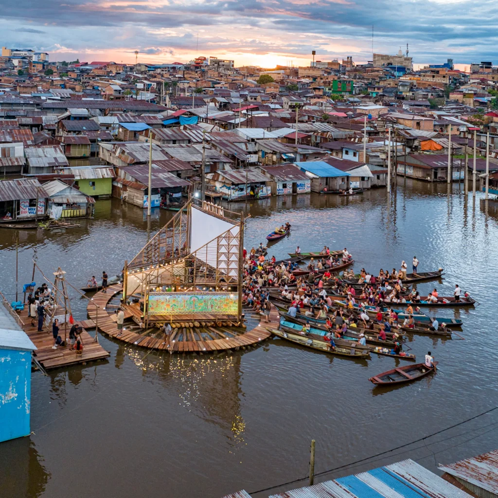 Floating wooden stage and seating platform on a river at sunset, surrounded by boats and dense riverside settlement