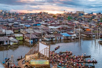 Floating wooden stage and seating platform on a river at sunset, surrounded by boats and dense riverside settlement