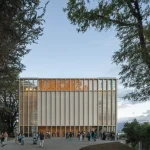 Rectangular timber-and-glass cultural building glowing at dusk with people gathered on the plaza in front, framed by large trees