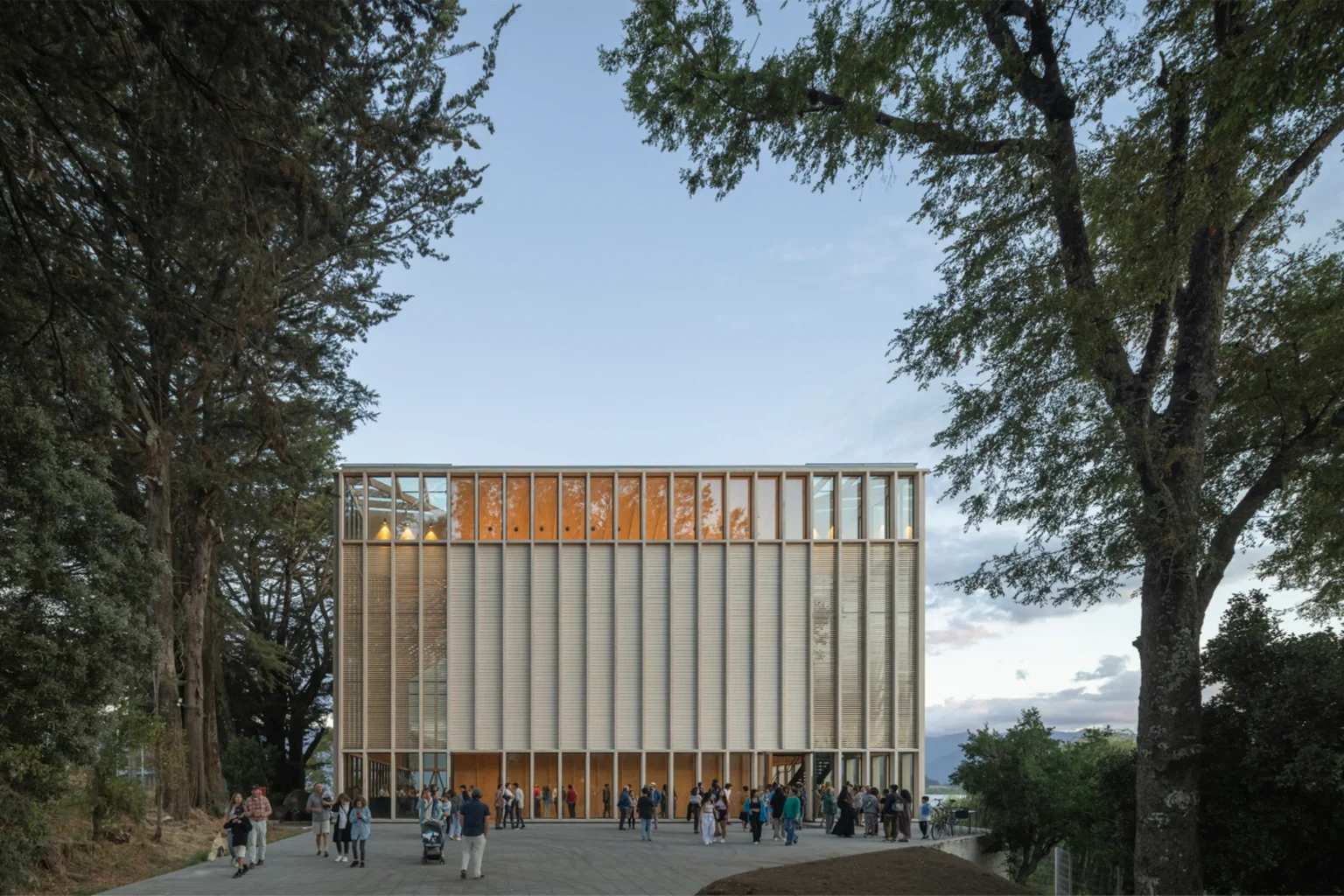 Rectangular timber-and-glass cultural building glowing at dusk with people gathered on the plaza in front, framed by large trees