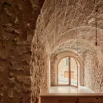 Vaulted stone interior with rough textured walls and an open arched doorway looking onto a sunlit street