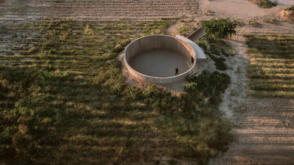 Circular concrete open-air structure set in dry agricultural fields with a single figure standing inside