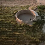 Circular concrete open-air structure set in dry agricultural fields with a single figure standing inside