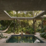 Covered concrete outdoor room with a small pool, hammock and dense tropical planting, a person seated at the water’s edge