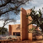 Earth-toned concrete chapel structure standing among dry trees and cacti in an arid landscape