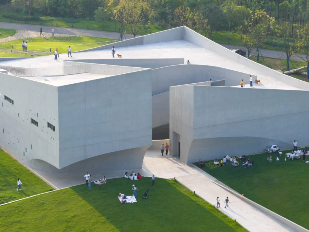Close aerial view of the multi-faceted concrete museum with people standing on the roof terraces and others picnicking on the grass below