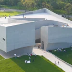 Close aerial view of the multi-faceted concrete museum with people standing on the roof terraces and others picnicking on the grass below