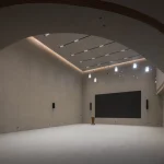 Interior of a large concrete hall with curved ceiling, soft lighting and a dark projection screen at the far end