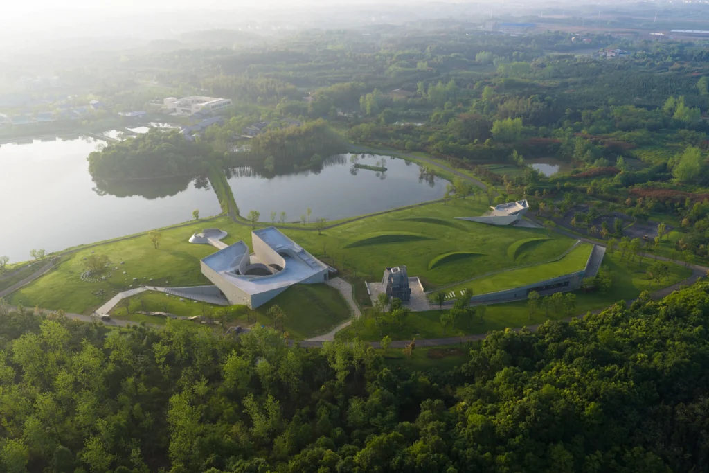 Aerial view of the Zi Museum cluster set in green meadows beside a lake, with the main angular concrete building, meditation tower and grass-covered volumes visible at sunrise