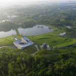 Aerial view of the Zi Museum cluster set in green meadows beside a lake, with the main angular concrete building, meditation tower and grass-covered volumes visible at sunrise