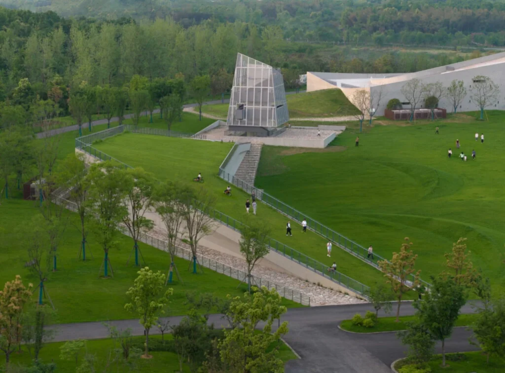 View of the glass meditation tower and long ramped pathways cutting through the green landscape with people walking and cycling
