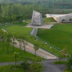 View of the glass meditation tower and long ramped pathways cutting through the green landscape with people walking and cycling