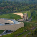 Elevated view of the main concrete museum building with curved openings and the glass meditation tower standing on a raised grass platform